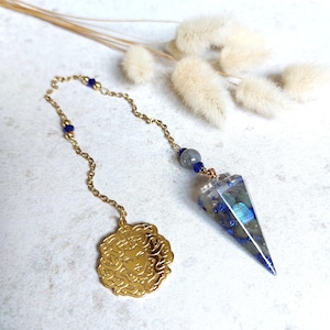 Puede incluir: Un péndulo de cadena de metal dorado con un colgante de resina puntiagudo incrustado con piedras azules y grises. La cadena incluye pequeñas cuentas azules y termina en un dije redondo dorado con un rostro detallado rodeado de serpientes.