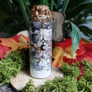 Peut inclure: Une bouteille en verre transparente remplie de couches de cristaux, de coquillages et de sable. La bouteille est scellée avec un bouchon en liège et de la cire dorée. Un charme avec un croissant de lune et une étoile est suspendu à la bouteille. L'étiquette indique "Améthyste, Quartz & Lavande."