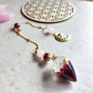 May include: A handcrafted pendant with a clear resin teardrop filled with red and gold details. The pendant is suspended from a gold chain with pink and red beads, and a gold crescent moon charm.