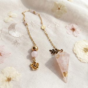 Könnte beinhalten: Ein Pendel mit goldfarbener Metallkette und einer facettierten, durchscheinenden rosa Steinspitze mit Blattgoldeinschlüssen. Die Kette ist mit einem kleinen goldenen Blumenanhänger, einer runden rosa Perle und einem blattförmigen Verbindungsstück verziert.
