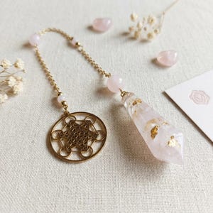 Op de afbeelding: Een pendel met een heldere, gefacetteerde kristallen punt met goudkleurige vlokken erin, bevestigd aan een goudkleurige ketting met lichtroze kralen en een ronde, goudkleurige metalen bedel met een geometrisch sterpatroon.