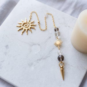 Puede incluir: Un péndulo de metal dorado con un dije en forma de sol en un extremo, una cadena delicada y una secuencia vertical de cuentas esféricas claras y oscuras intercaladas con un pequeño dije de sol, que termina en una punta de metal dorado.