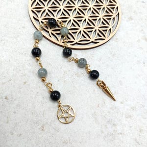 Könnte beinhalten: Goldfarbene Ohrring-Sets mit schwarzen und grauen Perlen, einem Pentagramm-Anhänger und einem goldenen Stachel. Die Ohrringe werden auf einer hellen Oberfläche mit einem dekorativen Holzelement im Hintergrund präsentiert.