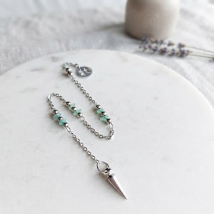 Könnte beinhalten: Ein silberfarbenes Metallpendel mit einer spitzen Form, das an einer feinen Kette befestigt ist. Die Kette ist mit kleinen, polierten Silberperlen und mehreren hellgrünen, rohen Steinsplittern verziert. Am Ende befindet sich ein kleiner runder Metallanhänger.