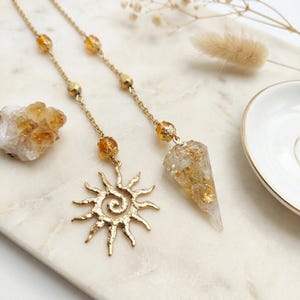 Op de afbeelding: Een goudkleurige ketting met twee verschillende hangers: een gestructureerde zon-bedel met een spiraalvormig midden en een conische hanger van heldere hars gevuld met goudvlokken. Beide kettingen zijn versierd met kleine barnsteenkleurige kralen.