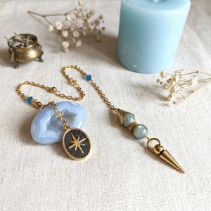 May include: A gold-toned chain necklace featuring a blue agate geode pendant with a starburst emblem, paired with a second chain segment ending in a pointed metal tip and two round grey beads, accented with small blue beads along the chain links.