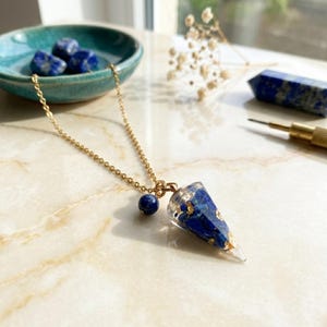 Könnte beinhalten: Eine goldfarbene Kettenhalskette mit einem konischen Anhänger aus klarem Harz, in den blaue Lapislazuli-Fragmente und Blattgoldflocken eingearbeitet sind. Eine kleine, runde Lapislazuli-Perle ist am Verbindungsring nahe der Anhängerspitze befestigt.