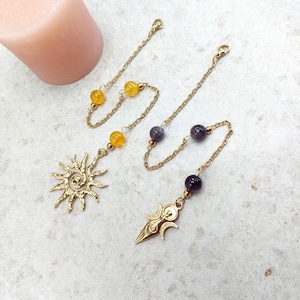 May include: Gold-coloured jewellery pieces with chain and bead accents. One features a sunburst pendant, the other a crescent moon design. The chains are adorned with yellow and purple beads.