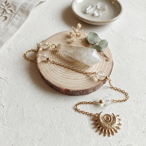 Könnte beinhalten: Eine goldfarbene Metallkette mit einem klaren, facettierten Kristallanhänger und einem sonnenförmigen Anhänger mit einer zentralen Spirale. Die Kette ist mit kleinen, klaren, kugelförmigen Perlen verziert und liegt auf einer runden Holzscheibe.