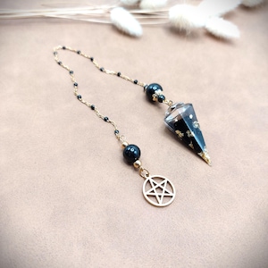 Peut inclure: Un pendule avec une chaîne dorée et des perles noires. Le pendule a une forme conique transparente et facettée avec des accents noirs et dorés. Un charme de pentagramme doré est suspendu à la chaîne.