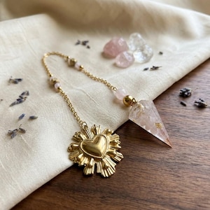 May include: A gold-toned metal chain features a pointed, translucent crystal pendant with gold flecks and a gold-toned heart-shaped charm with radiating rays. The chain is accented with small, pale pink beads, creating a decorative hanging ornament.