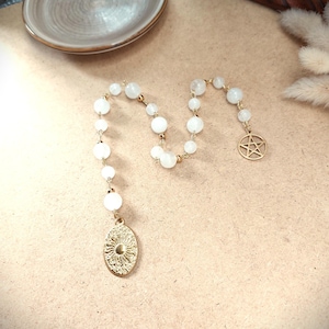 Könnte beinhalten: Eine goldfarbene Halskette im Rosenkranz-Stil mit weißen, runden Perlen. Die Halskette hat einen goldenen Sonnenanhänger und einen goldenen Pentagramm-Anhänger. Die Perlen sind auf einer goldenen Kette aufgereiht.
