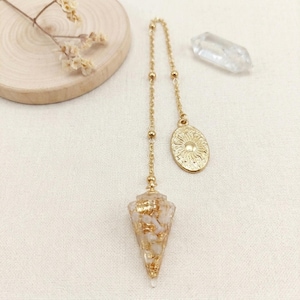 Könnte beinhalten: Ein goldfarbenes Pendel mit einem konischen Anhänger, der mit weißen Steinsplittern und Goldflocken gefüllt ist und an einer feinen Kette hängt. Ein ovaler Anhänger mit eingeprägtem Sonnenmotiv ist an der Kette befestigt, ergänzt durch einen klaren Kristall.