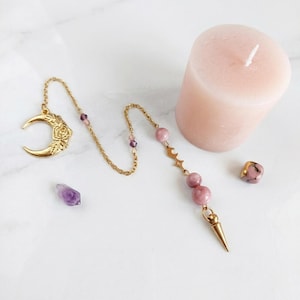 Könnte beinhalten: Ein goldfarbenes Metallpendel mit einem Mondsichel-Anhänger mit Blumenmuster, verbunden durch eine feine Kette mit zwei runden rosa Perlen und einer spitzen Metallspitze. Eine blassrosa Stumpenkerze und zwei kleine lose Kristalle liegen daneben.