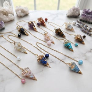 Könnte beinhalten: Eine Sammlung von kegelförmigen Pendeln aus Kunstharz mit eingebetteten Steinen und goldfarbenen Flocken. Jedes Pendel hängt an einer goldfarbenen Metallkette, die mit einer kleinen runden Perle in verschiedenen Farben wie Blau, Rosa und Weiß endet.