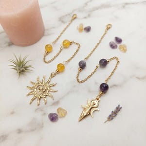 Könnte beinhalten: Zwei goldfarbene Metallketten mit Edelsteinperlen. Eine Kette hat gelbe Perlen und einen sonnenförmigen Anhänger, die andere violette Perlen und einen dreifachen Mondanhänger. Lose gelbe und violette Edelsteinperlen liegen daneben.