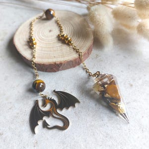 Peut inclure: Un pendule avec une chaîne dorée, des perles en œil de tigre et un charm en forme de dragon. Le pendule a un corps en résine transparente rempli de pierres en œil de tigre et de paillettes dorées. Le charm dragon est marron foncé avec des accents orange.