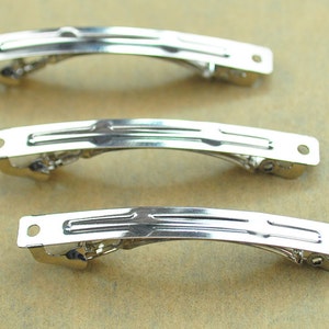 Peut inclure: Trois barrettes à cheveux en métal argenté. Chaque barrette a une forme rectangulaire incurvée avec un fermoir à charnière et un petit trou à chaque extrémité. Les barrettes sont présentées sur une surface brun clair.