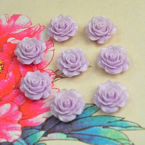 Könnte beinhalten: Acht rosafarbene Harz-Rosen-Cabochons, jeder mit einer leicht unterschiedlichen Form und Größe, sind auf einem floralen Hintergrund angeordnet.