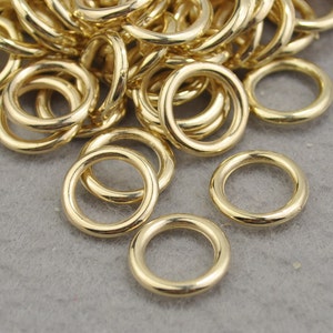 May include: A collection of shiny, gold-colored jump rings. The rings are circular, with a smooth, polished surface. They are scattered on a gray surface, with some rings overlapping. The lighting highlights the metallic sheen of the rings.