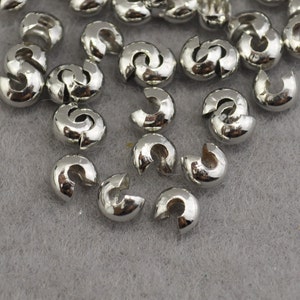 May include: A close-up of a pile of silver-toned metal crimp beads. The beads are small and round, with a hole in the centre. They are used for jewellery making.