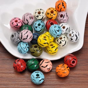May include: A collection of colorful wooden beads with a round shape and a hole through the center. The beads are painted in various colors, including pink, orange, yellow, and blue, with black curved patterns. They are displayed on a white plate.