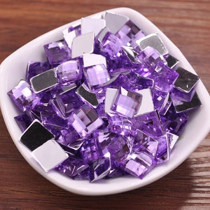 May include: A large assortment of purple and clear square-shaped rhinestones. The rhinestones are scattered in a white bowl.