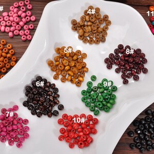 May include: A selection of wooden beads in a range of colours, such as pink, orange, yellow, brown, green, red, and black. The beads are displayed in small piles on a white, leaf-shaped dish. The image is well-lit, highlighting the colours and textures of the beads.