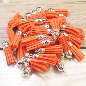 May include: A large group of orange tassel charms with silver metal caps. The tassels are made of soft, fuzzy material and are perfect for adding a touch of color and texture to crafts.