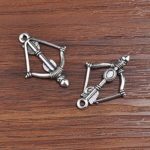 Puede incluir: Dos abalorios de arco y flecha plateados con detalles intrincados. Los abalorios tienen un bucle en la parte superior para sujetarlos a joyas u otras manualidades.