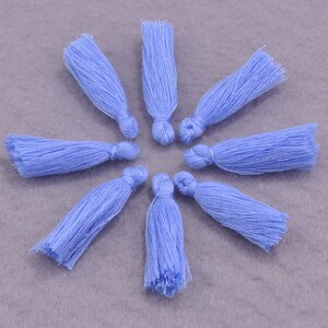 Puede incluir: Ocho borlas de color azul claro dispuestas en un patrón circular. Cada borla está hecha de hilos bien enrollados, con una parte superior anudada y una parte inferior con flecos. Las borlas son de un color azul pastel suave.