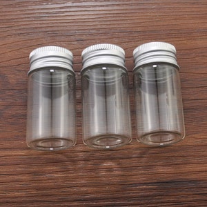May include: Three small, clear glass jars with silver metal screw-on lids. The jars are cylindrical and empty, suitable for storing small items. The lids have a ridged design for easy gripping.