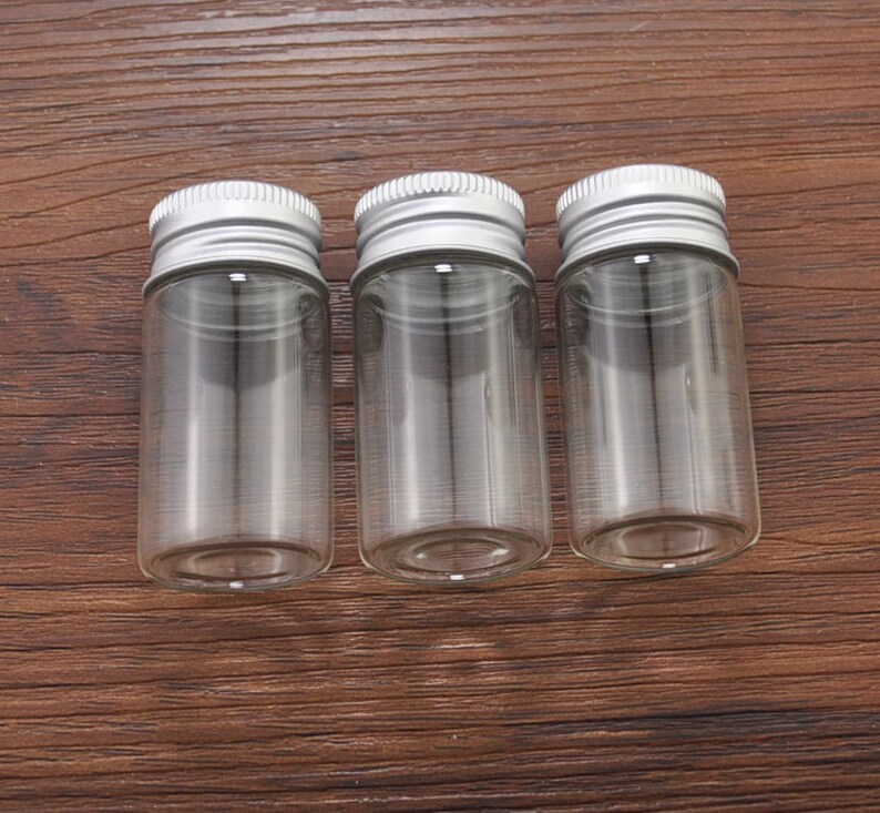 May include: Three small, clear glass jars with silver metal screw-on lids. The jars are cylindrical and empty, suitable for storing small items. The lids have a ridged design for easy gripping.