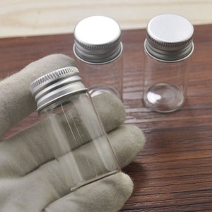May include: Clear glass vials with silver metal screw-on lids. One vial is held in a gray gloved hand, with two other vials in the background. The vials are cylindrical and appear empty. The background is a wooden surface.