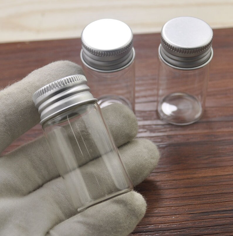 May include: Clear glass vials with silver metal screw-on lids. One vial is held in a gray gloved hand, with two other vials in the background. The vials are cylindrical and appear empty. The background is a wooden surface.