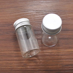 May include: Two small, clear glass bottles with silver metal screw-on lids. The bottles are cylindrical and empty, resting on a dark brown wooden surface. One bottle is lying on its side, while the other stands upright.