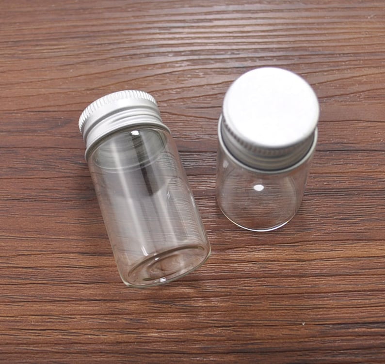 May include: Two small, clear glass bottles with silver metal screw-on lids. The bottles are cylindrical and empty, resting on a dark brown wooden surface. One bottle is lying on its side, while the other stands upright.