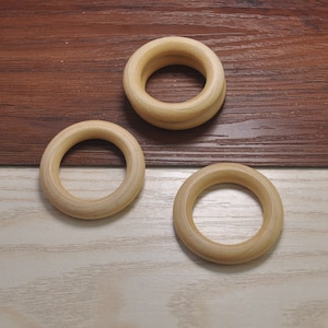 May include: Three wooden rings of different sizes, all with a natural wood finish. The rings are arranged on a light brown surface.