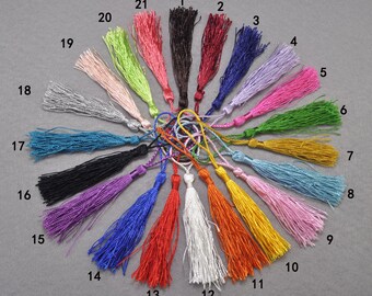 May include: A collection of 21 colorful tassels, each with a looped top and a long, fringed body. The tassels vary in color, including black, red, blue, purple, green, yellow, orange, pink, and silver. They are arranged in a circular pattern against a gray background.