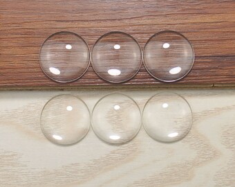 May include: Six clear glass cabochons, round and smooth, arranged in two rows of three on a wooden and white surface.