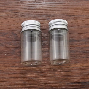 Op de afbeelding: Twee kleine, heldere glazen flesjes met zilveren metalen schroefdoppen. De flesjes zijn cilindrisch en staan op een donkerbruin houten oppervlak. De doppen hebben een geribbelde textuur. De flesjes zijn leeg.