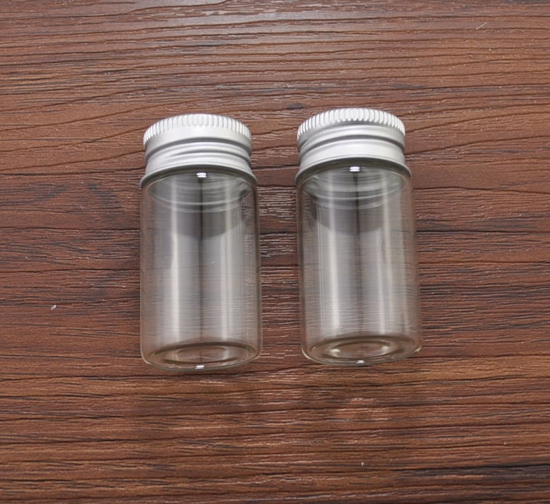 May include: Two small, clear glass bottles with silver metal screw-on lids. The bottles are cylindrical and sit on a dark brown wooden surface. The lids have a ridged texture. The bottles are empty.