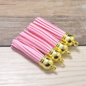 May include: Four pink suede tassels with gold metal caps and loops. The tassels are arranged in a row, with the caps facing the viewer.