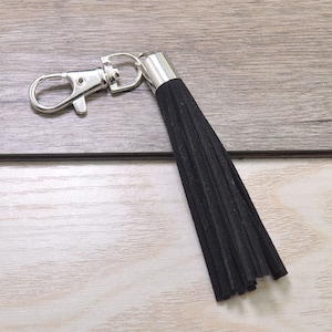 Puede incluir: Un llavero de borla negro con un anillo y un clip de metal plateado.