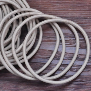 May include: A pile of light gray elastic hair ties, also known as scrunchies, are arranged on a dark wood surface. The hair ties are circular and made of a soft, fabric-covered elastic material.