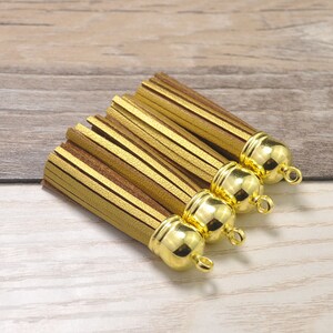 May include: Four gold and brown tassels with gold-colored caps and small loops. The tassels are made of layered strips of material, with a mix of gold and brown colors. The background is a light brown wooden surface.