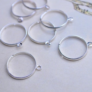 May include: A set of six silver-toned metal rings with a jump ring attached to each. The rings are all the same size and are arranged in a random pattern on a white surface.