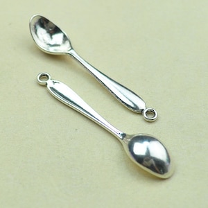 May include: Two silver spoons with a rounded bowl and a loop at the top of the handle. The spoons are on a light beige surface.