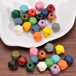 May include: A collection of colorful, geometric wooden beads in various shades, including pink, blue, green, yellow, and orange. The beads are displayed on a white, scalloped-edge dish and scattered on a wooden surface.