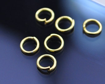 May include: Close-up of several small, gold-colored jump rings. The rings are open, with a small gap in the circle. The background is a dark, reflective surface. These jewelry findings are used for connecting jewelry components.